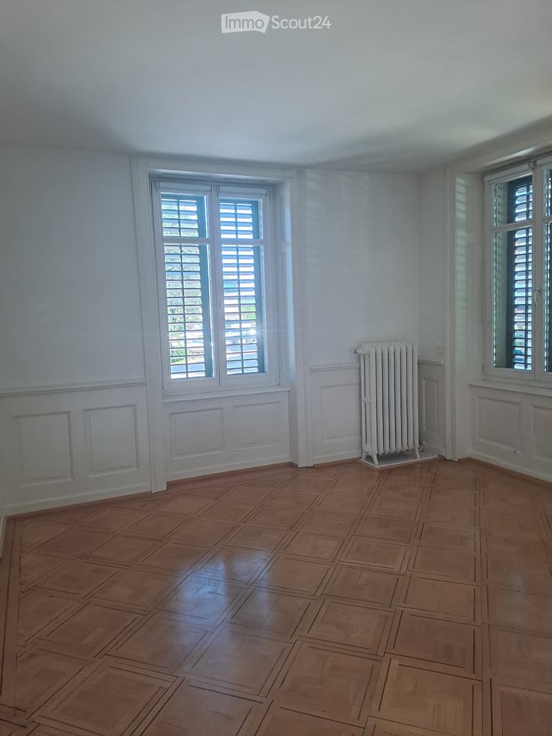 Empty room with wooden parquet flooring, white walls, and two windows with shutters. A white radiator is placed against one wall.