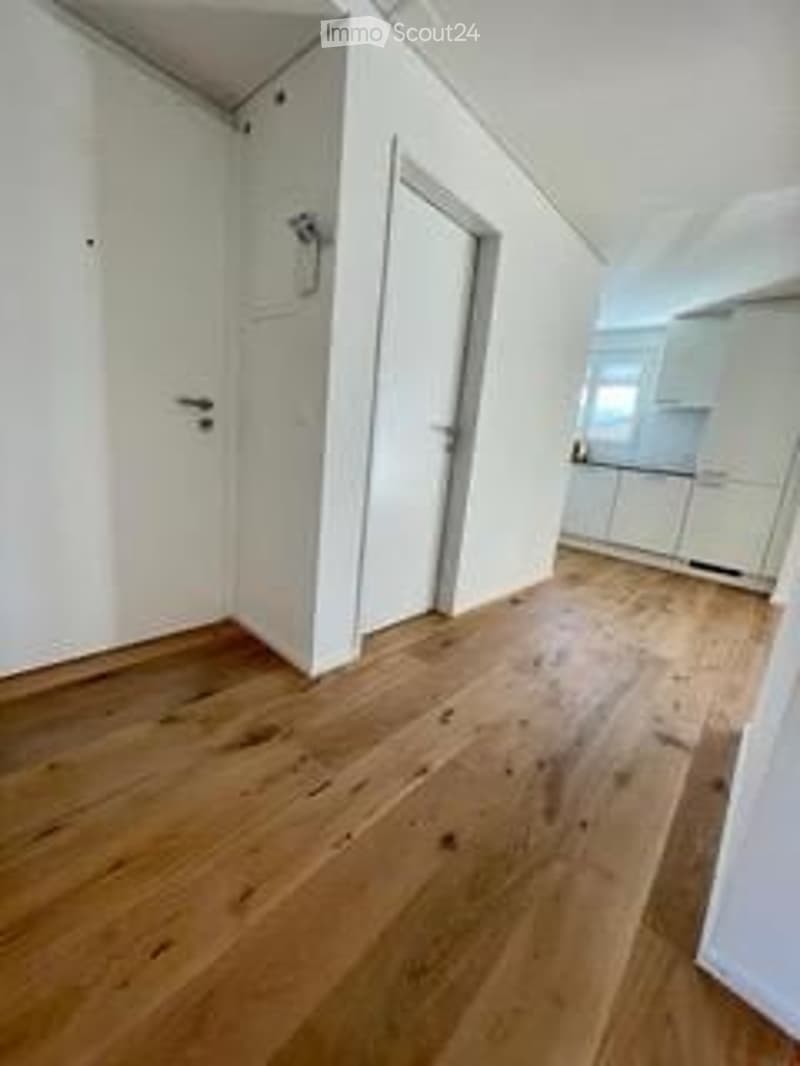 Completely empty room with white walls, wooden floor, and a door leading to another room.