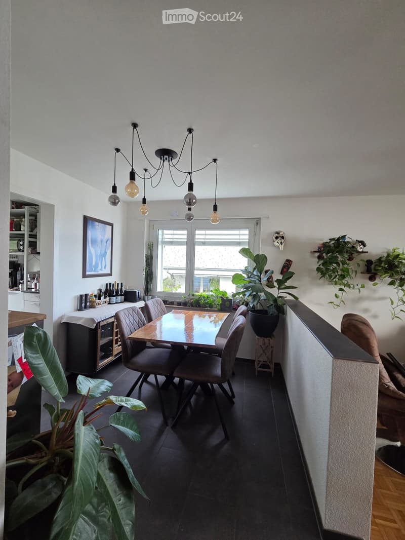 Dining area with a wooden table, six chairs, large potted plants, and a cabinet with bottles and a coffee maker. A chandelier hangs from the ceiling, and a window offers natural light.