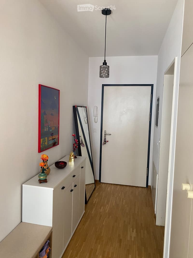 A narrow hallway with wooden flooring, white walls, a hanging light fixture, a white cabinet with decorative items, a mirror, and a door leading to another room.
