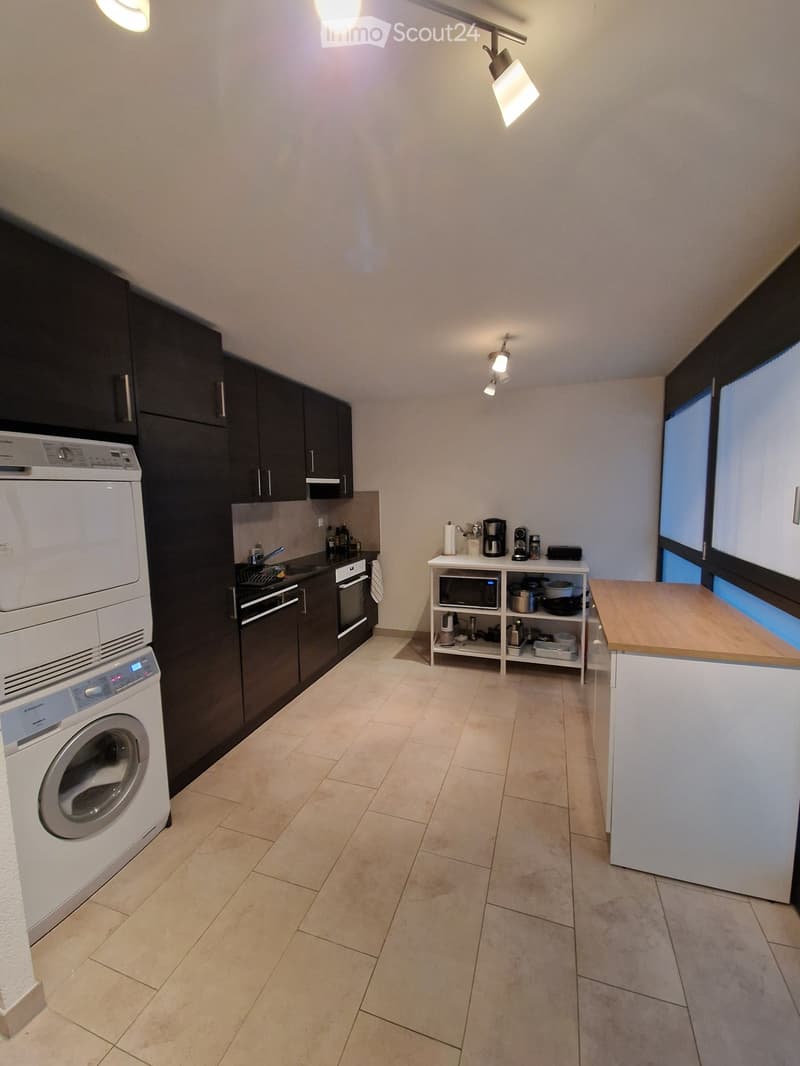 Modern kitchen with tiled floor, dark cabinets, white appliances including washing machine and oven, countertop with microwave and coffee maker, and a small shelf unit with various items.