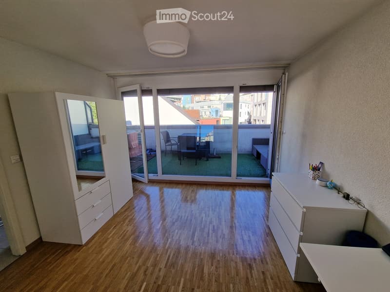 Bright room with wooden flooring, large sliding glass doors leading to a balcony, a wardrobe, a desk with office supplies, and a small seating area visible through the glass doors.