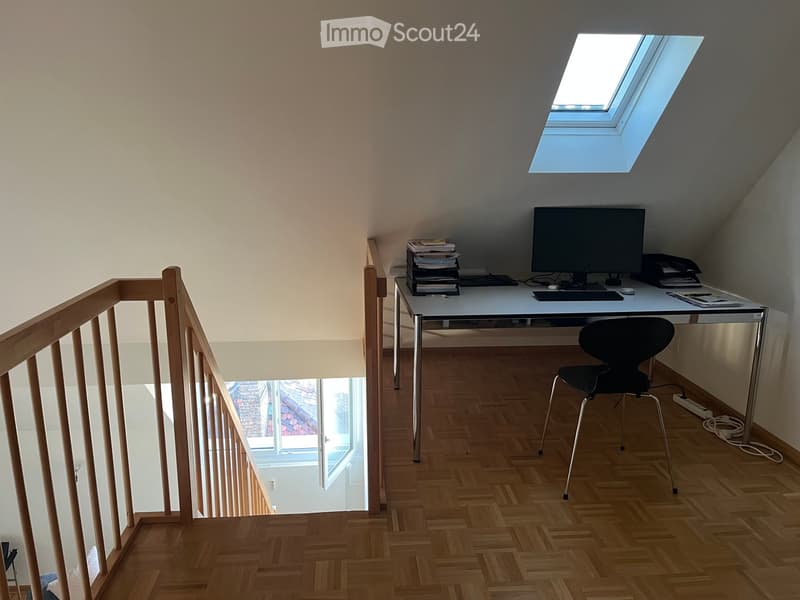 A modern, well-lit room with wooden flooring, a desk setup including a computer, chair, and various office supplies, and a staircase leading downwards.
