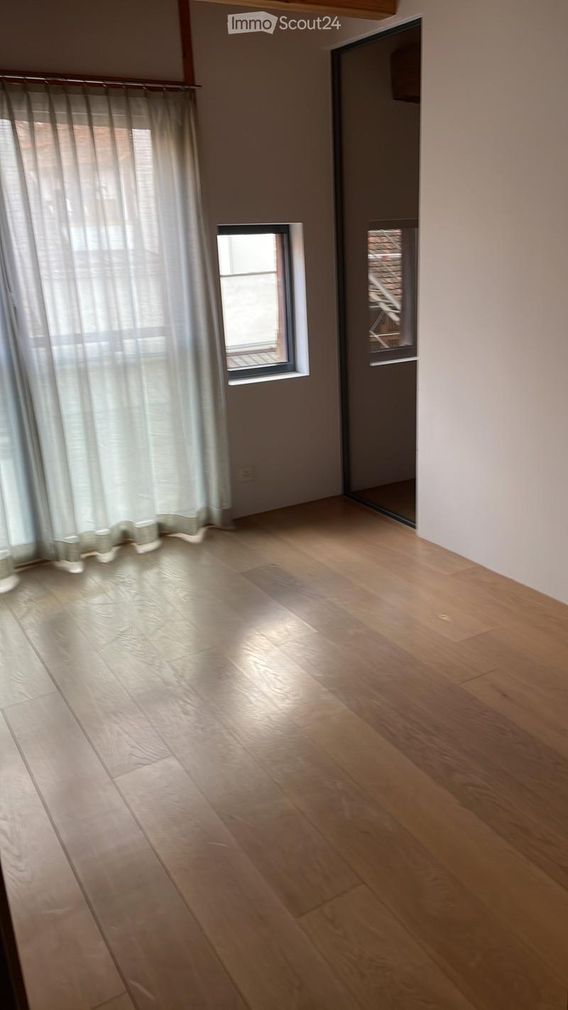 Empty room with wooden flooring, white walls, and a large window with sheer curtains. A small window and a doorway lead to another space.