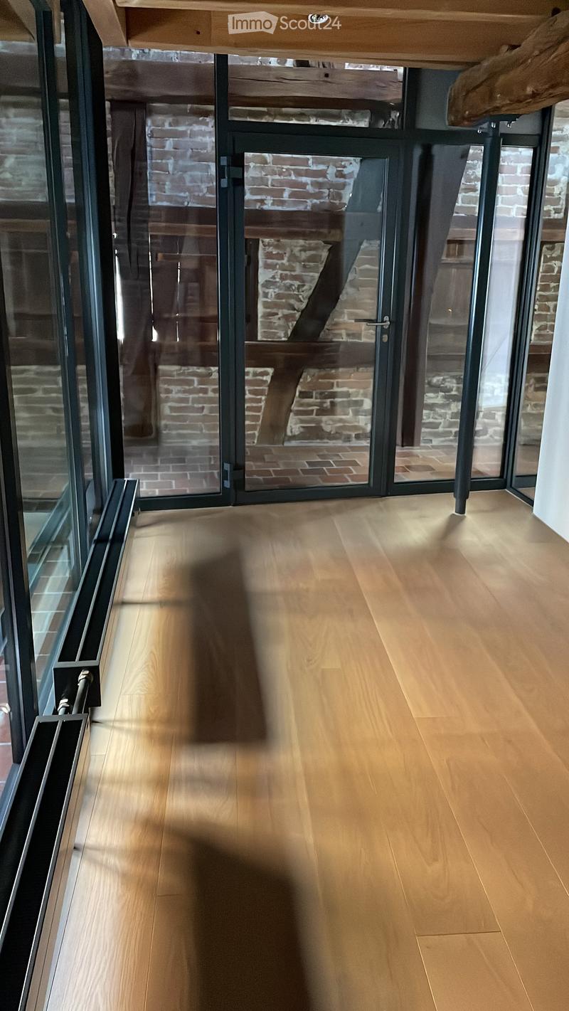Bright hallway with light wooden flooring, large glass doors leading to an outdoor area with brick walls and wooden beams visible through the glass.