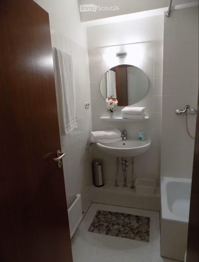 Modern bathroom with white tiles, a round mirror above the sink, neatly folded towels, a small flower vase, and a shower area.