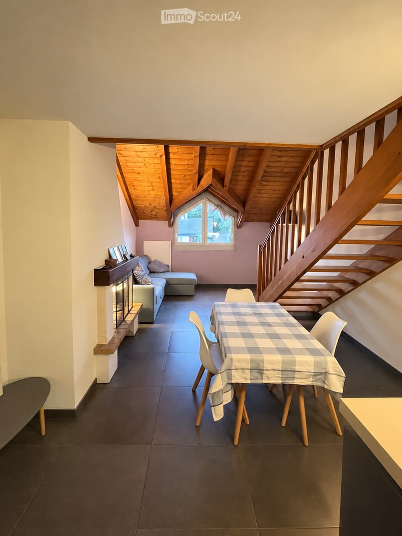 Bright living room with a wooden slanted ceiling, dark tiled floor, and a cozy seating area featuring a sofa, ottoman, and fireplace. A dining table with chairs is set with a checkered tablecloth, and a staircase leads upwards.