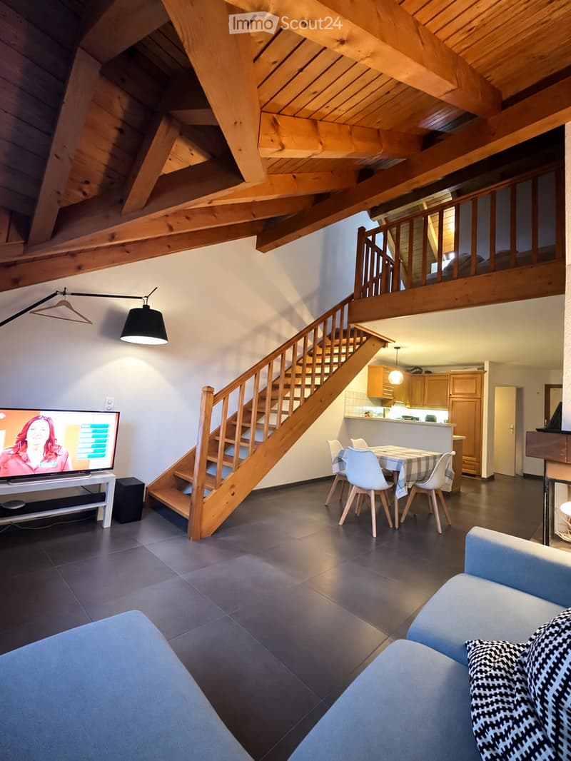 Open-plan living and dining area with a modern sofa, dining table with chairs, television, and a wooden staircase leading to an upper level. The kitchen area features light wooden cabinets and a countertop.