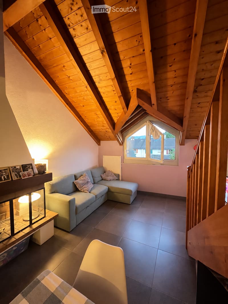 Cozy living room with a grey sectional sofa, fireplace, small table with lamp, and picture frames on a shelf. The room features wooden beams on the ceiling and a staircase leading upwards.