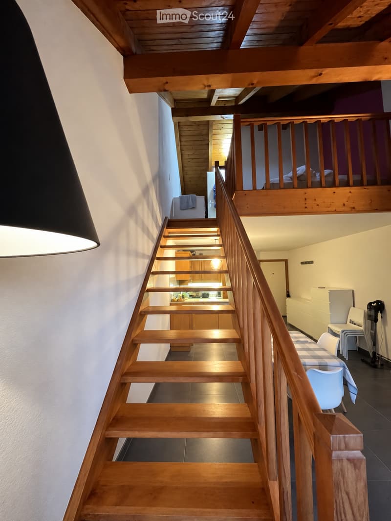 Wooden staircase with a wooden railing leads to an upper level. The lower level shows a kitchen area with cabinets and a dining area with a table and chairs.