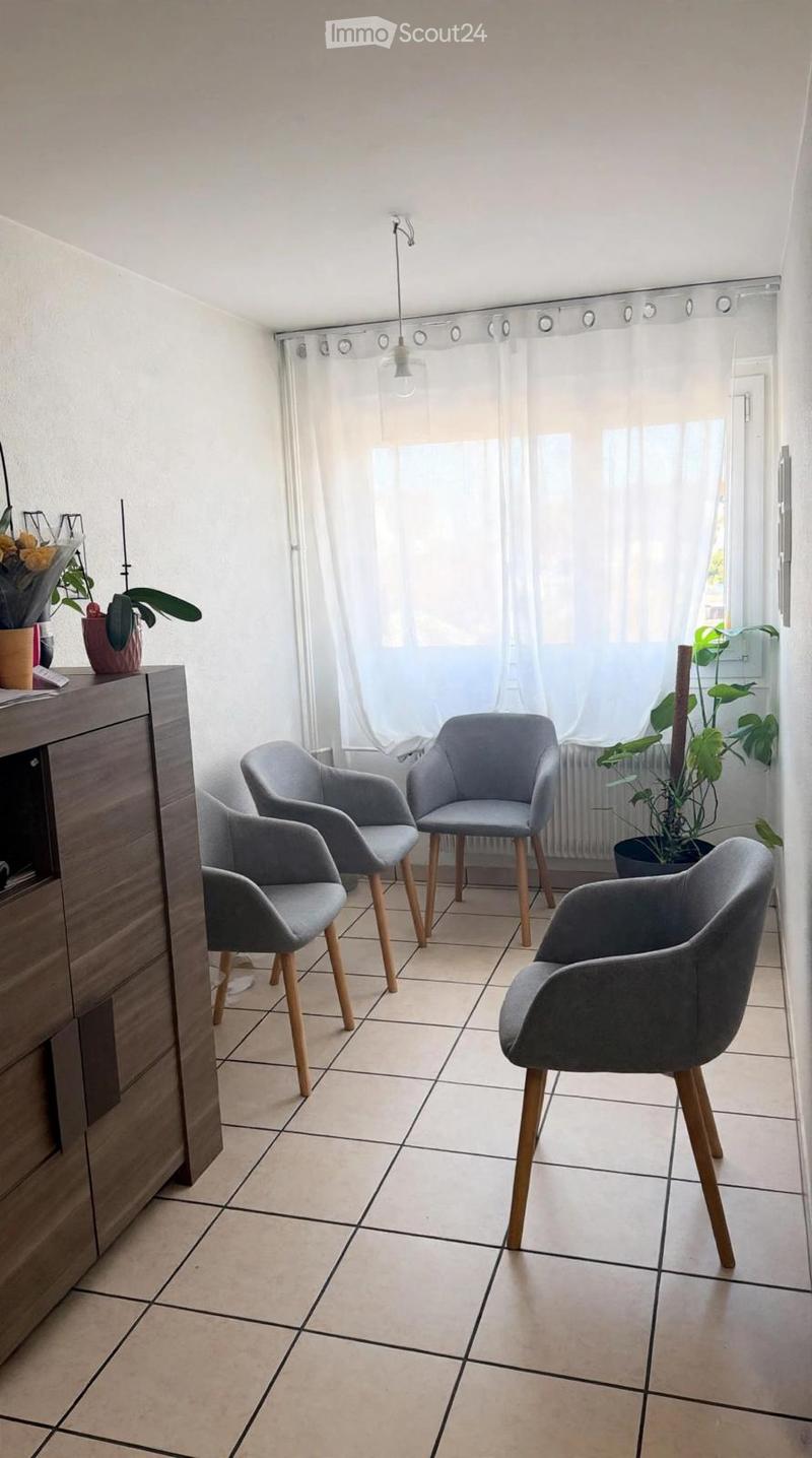 A small, well-lit room with tiled flooring and white walls. It features four modern grey chairs with wooden legs arranged in a semi-circle, a wooden cabinet with a drawer, and several potted plants near the window. The room has large windows covered with
