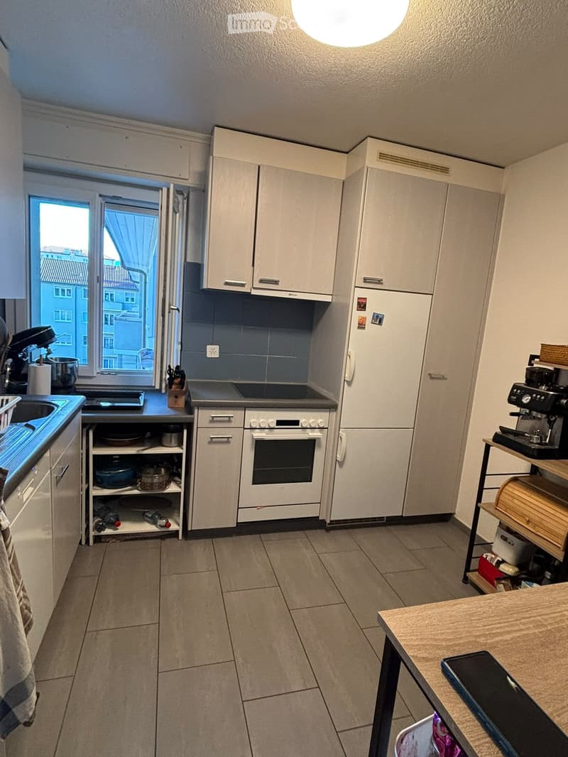 Modern kitchen with white cabinets, stainless steel appliances, and a large window offering a city view. Includes a sink, refrigerator, oven, coffee machine, and a small table.