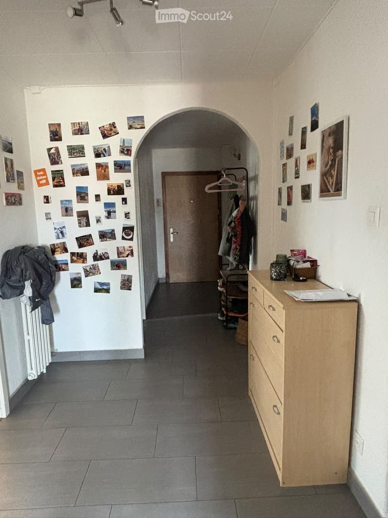 A narrow hallway with tiled flooring, white walls, and an arched doorway leading to another room. Photos are pinned to the left wall, and a small wooden cabinet with drawers is on the right side.