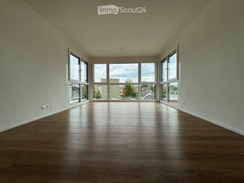 Empty room with wooden flooring and large windows offering a view of the outside. The walls are painted a light color, and the space is well-lit with natural light.