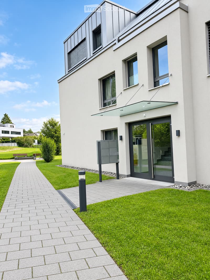 Modern two-story house with white facade, large windows, and a sleek entrance with glass doors. The surrounding area features a well-maintained lawn, a paved walkway, and a mailbox.