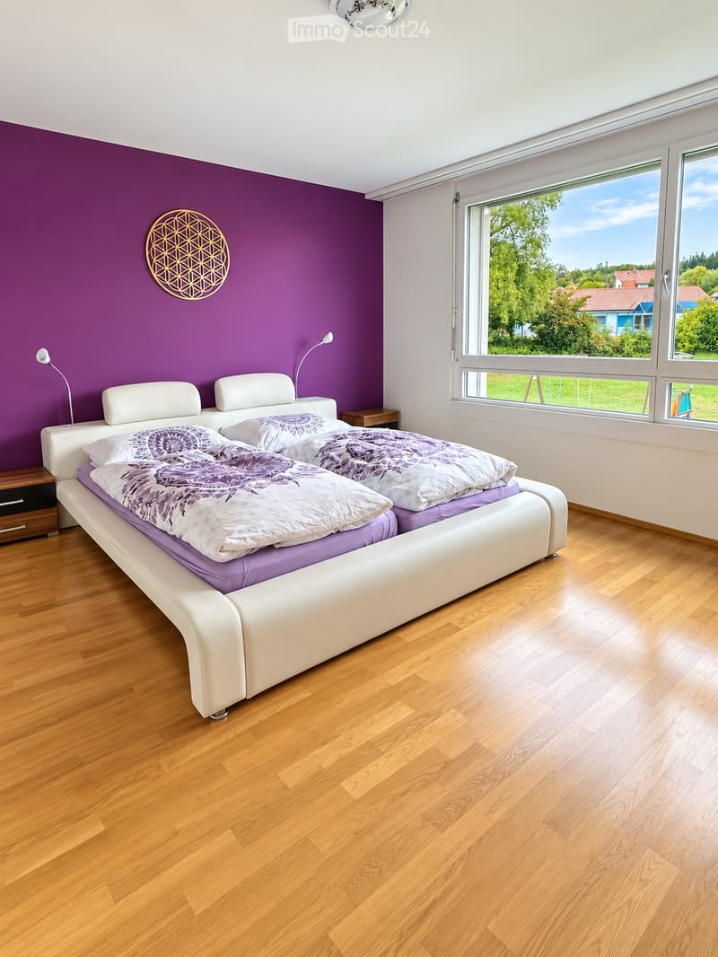 Modern bedroom with a large white leather bed, purple bedding, wooden nightstand, and wall-mounted lamp. Large windows offer a view of greenery outside.