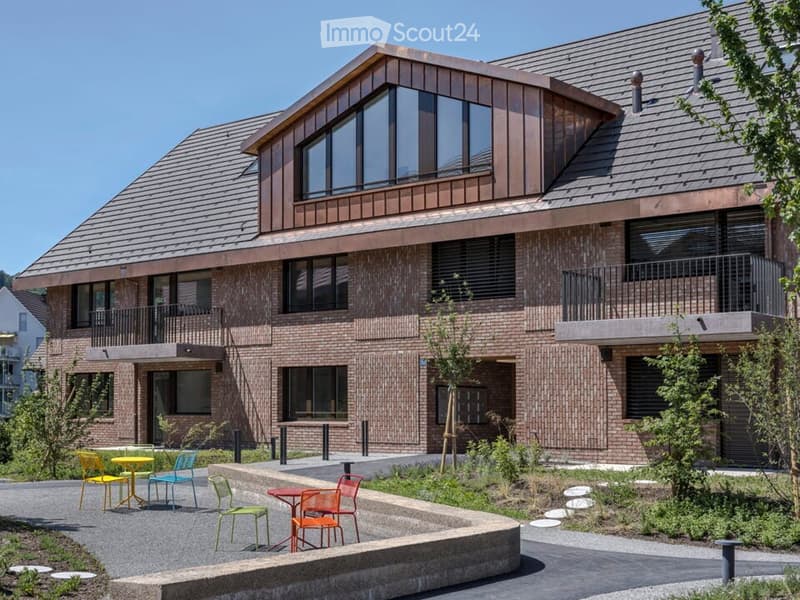 Modern two-story building with brick facade, large windows, and balconies. Outdoor seating area with tables and chairs is visible in front.