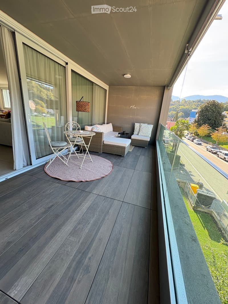 Balcony with wooden flooring, sofa, small table, chairs, and a view of the street and surrounding landscape.