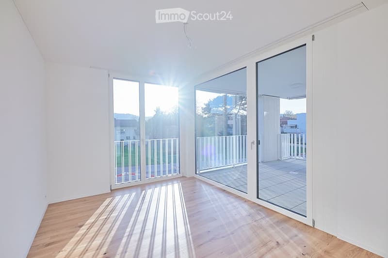 Bright, empty room with wooden flooring and large glass sliding doors leading to a balcony. Sunlight streams in, creating striped shadows on the floor.