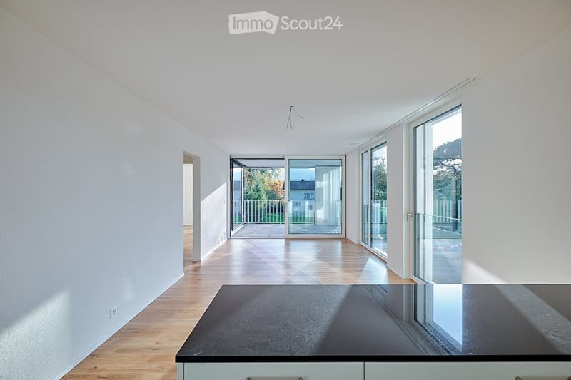 Bright, empty room with light wooden flooring, white walls, and large glass sliding doors leading to a balcony or outdoor area. The space is clean and minimalistic with no furniture or decorations.