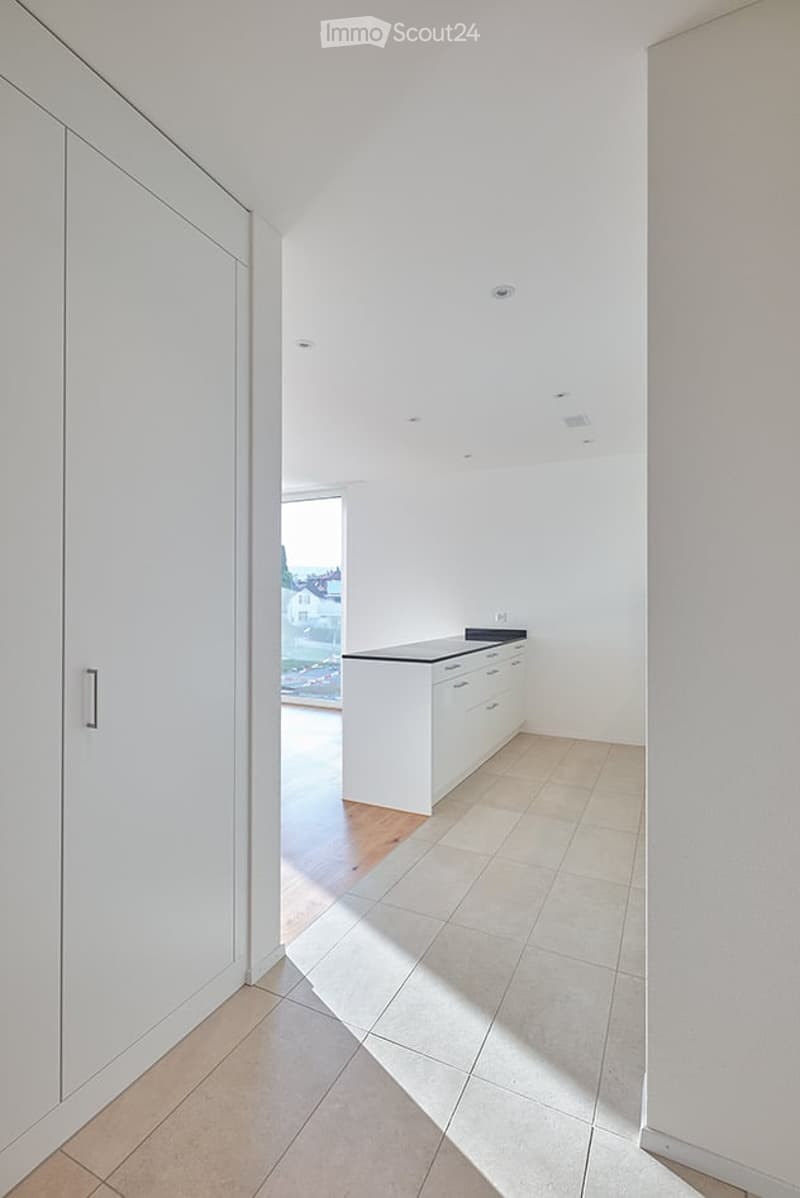 Empty room with white walls, tiled floor, and a small white kitchen unit visible in the background. No furniture or personal items present.
