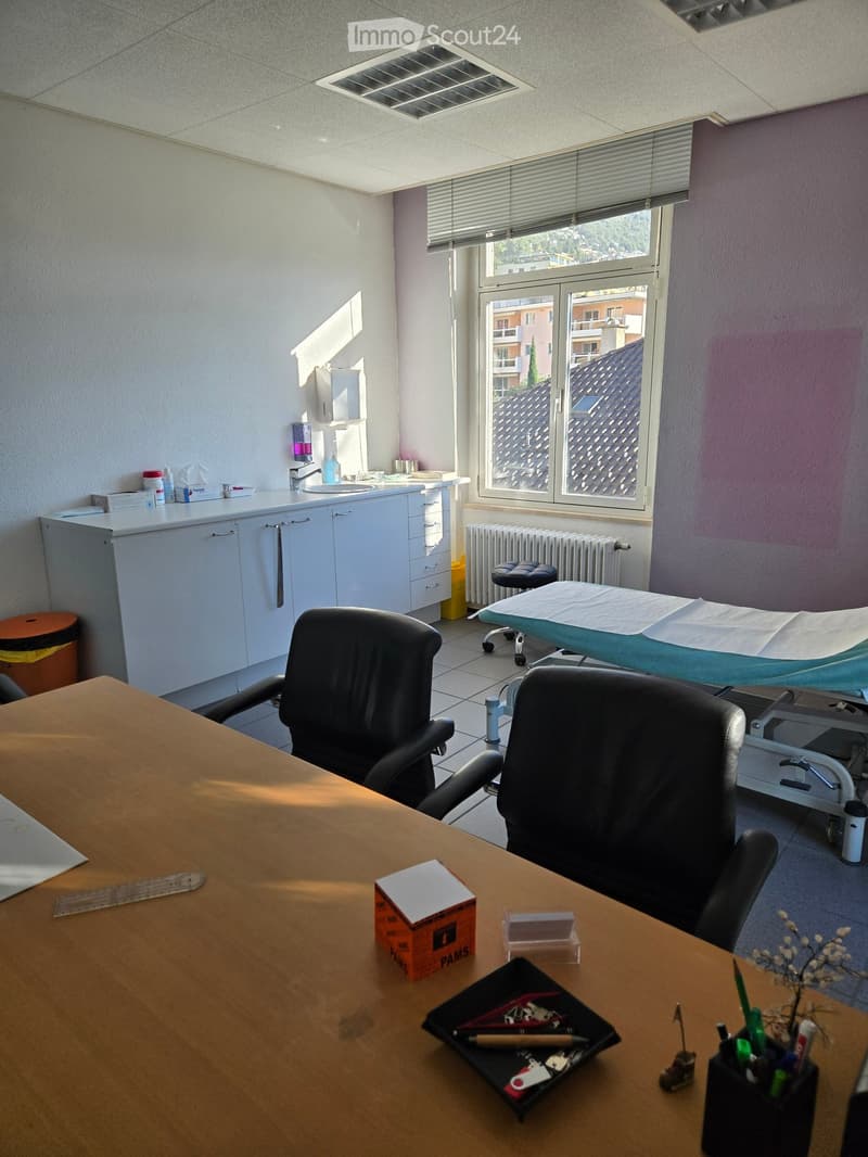 A well-lit room with a large window, a wooden table with chairs, a sink area with cabinets, an exam table, and a radiator. The room also includes a trash bin and a stool.