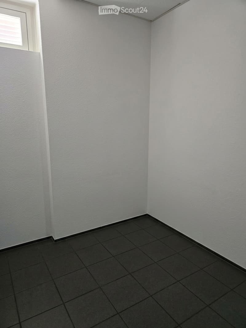 Empty room with white walls and dark tiled floor, featuring a small window with blinds on the left side.