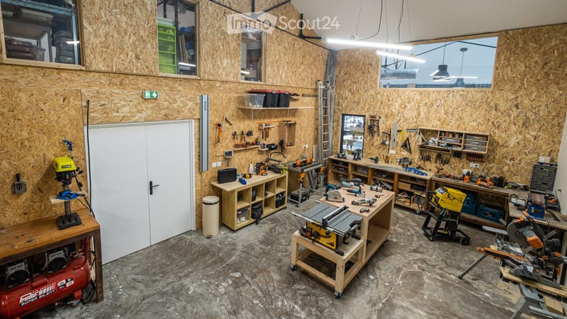 Well-organized workshop with wooden walls, concrete floor, and large windows. Contains workbenches, tools, storage shelves, and equipment like a table saw and air compressor.