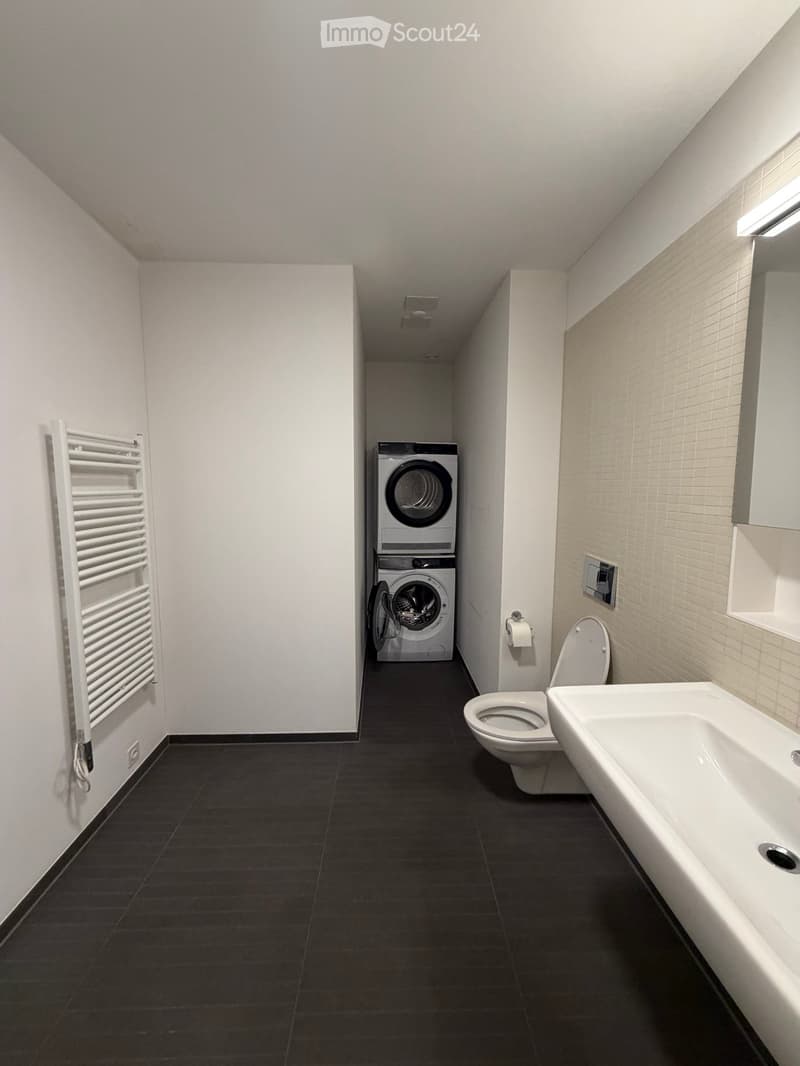 Modern bathroom with dark tiled floor, white radiator, toilet, sink, and stacked washing machine/dryer unit.