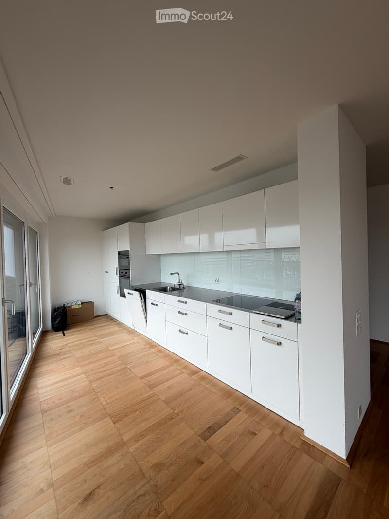 Modern kitchen with white cabinets, wooden flooring, and stainless steel appliances including a sink, stove, and dishwasher. A bottle is placed on the counter.