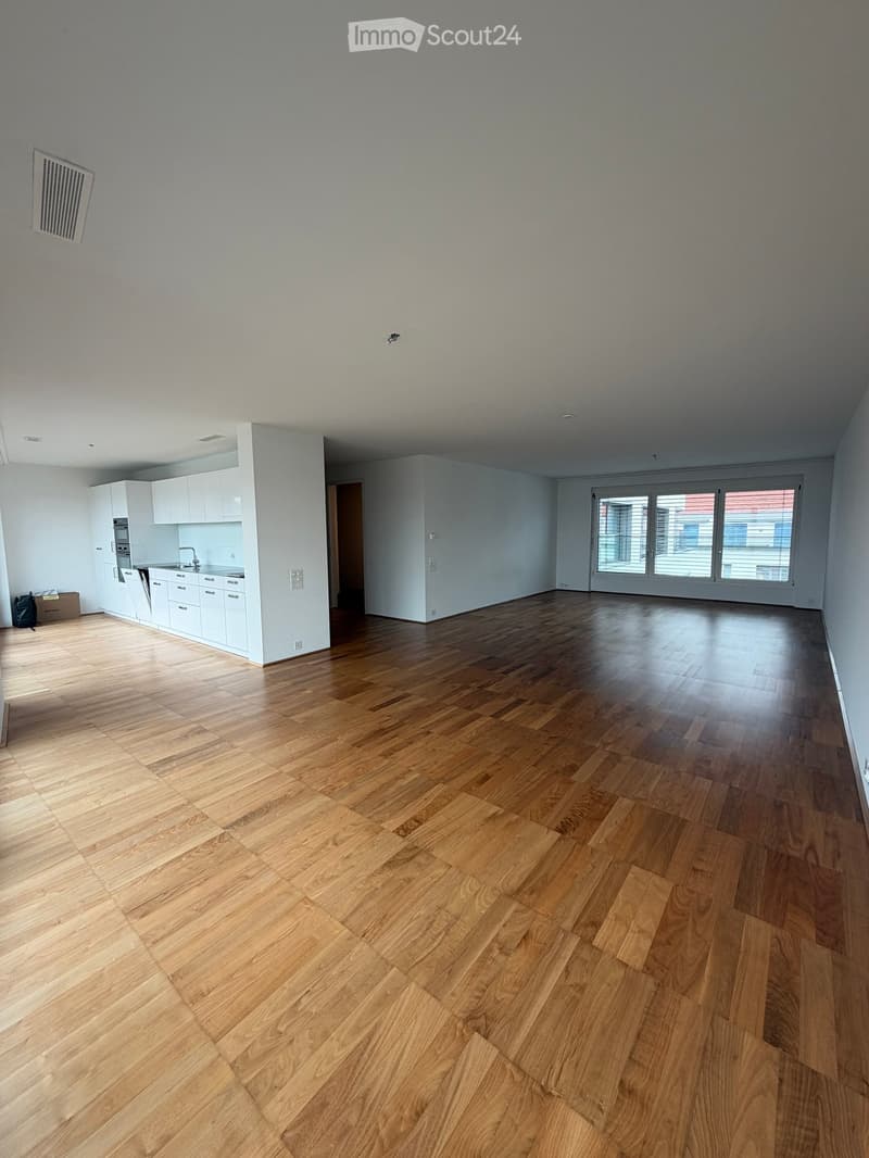 Empty room with light wooden flooring, white walls, and a small kitchen area with white cabinets and appliances visible in the background.