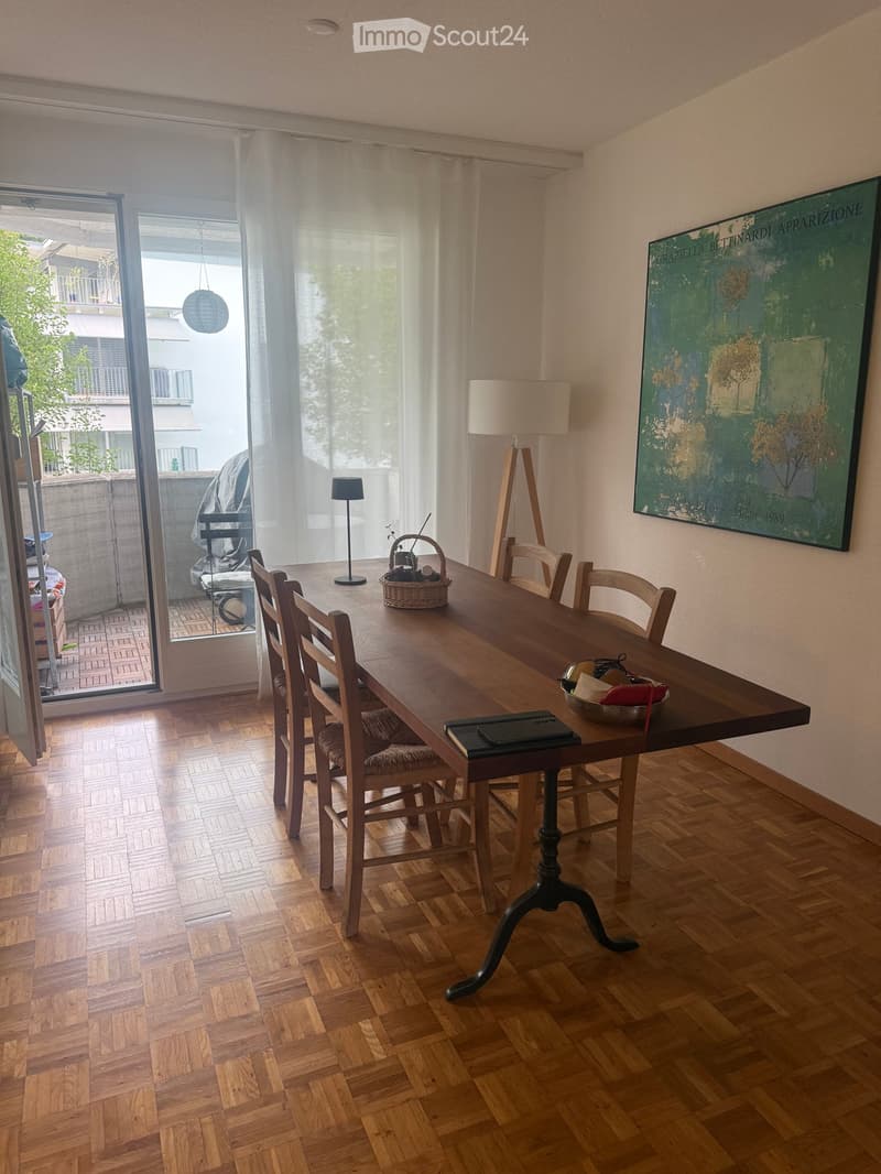 Bright kitchen with wooden herringbone flooring, large dining table surrounded by chairs, wicker basket, floor lamp, table lamp, and a large painting on the wall. Large window with sheer curtains and a view of a balcony.
