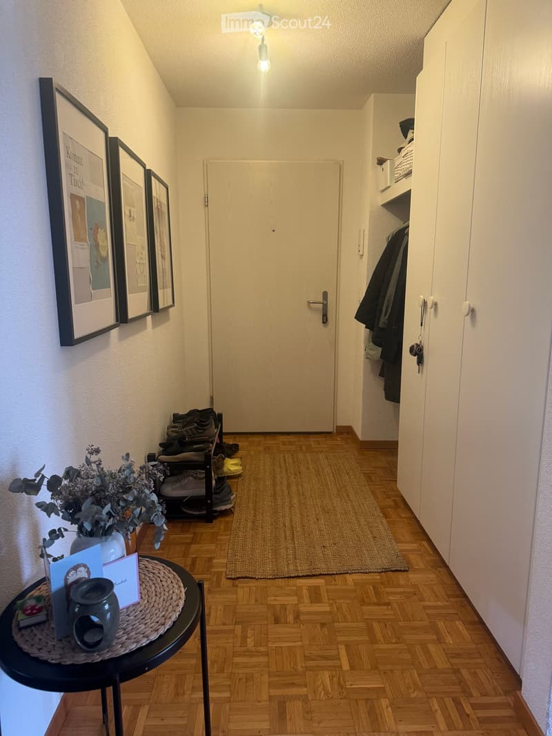 Narrow hallway with wooden flooring, a small round table with a candle and cards, a shoe rack with shoes, a coat closet, and framed artwork on the wall.