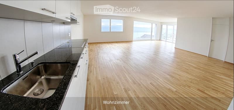 Empty room with wooden flooring, white walls, and large windows offering a sea view. Kitchen area with sink and cabinets visible on one side.