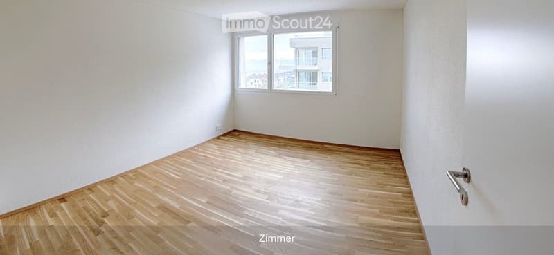 Empty room with light wooden flooring and white walls, featuring a large window with a view of a building and outdoor area.