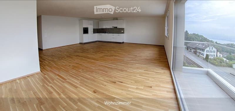 Open-plan living and dining area with wooden flooring, white kitchen cabinets, and large windows offering a scenic view.