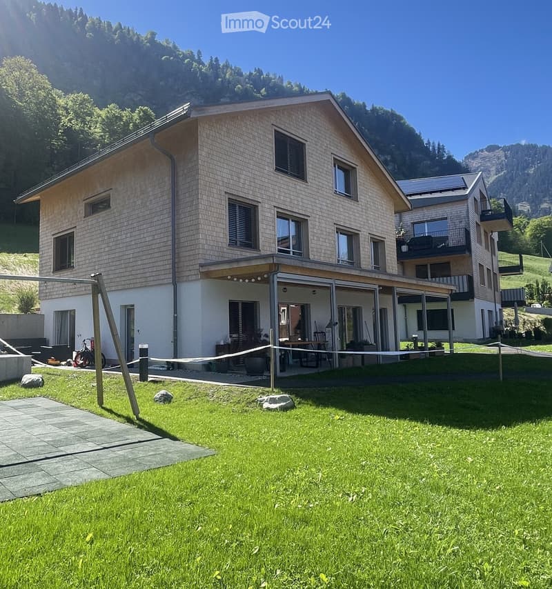 Two-story modern house with light-colored siding, large windows, and a spacious covered patio area. The house is surrounded by a well-maintained lawn and is situated in a scenic, mountainous area.