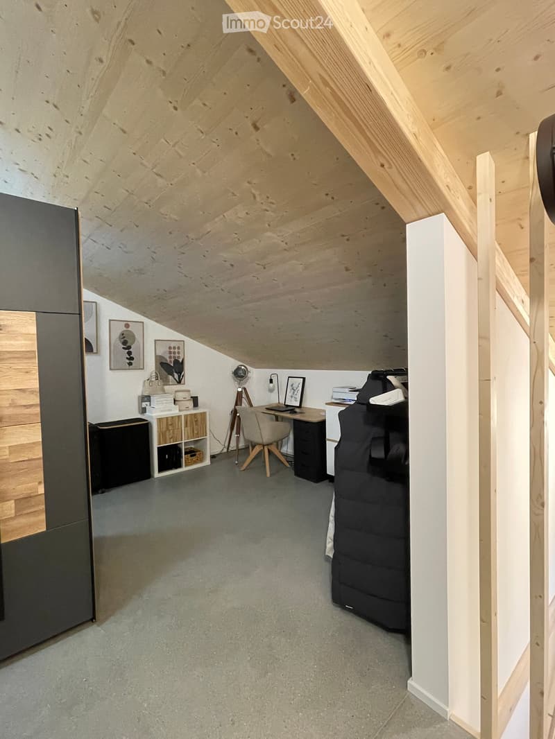 Loft-style room with exposed wooden beams, grey carpet, a sofa, desk, chair, and shelves with decorative items.