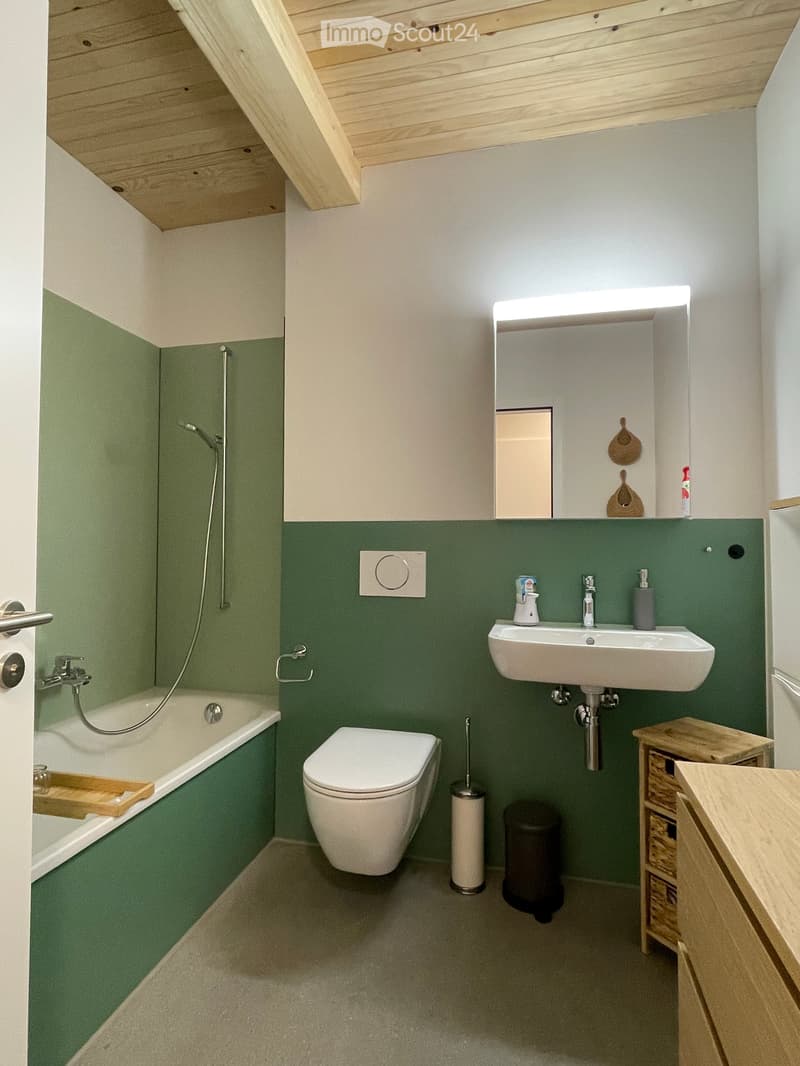 Modern bathroom with green lower walls, white upper walls, and a sloped wooden ceiling. Features a bathtub, toilet, sink with a mirror, and a small wooden stool with a woven basket.