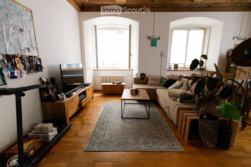Bright living room with wooden flooring, large windows, a sectional sofa, coffee table, TV, side table, and a plant in a pot.