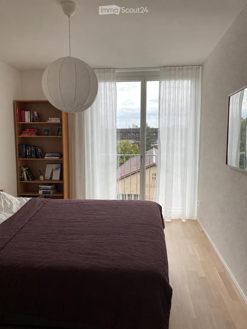 Modern bedroom with a dark brown duvet-covered bed, wooden floor, large window with sheer curtains, bookshelf, and a round pendant light.