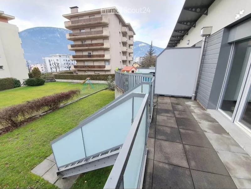 Balcony with modern glass railing, overlooking a green lawn and playground, with a multi-story residential building and mountain view in the background.