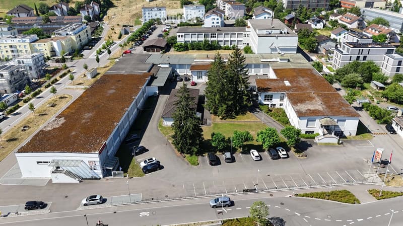 Aerial view of a commercial building with a large parking lot, surrounded by residential buildings and greenery.