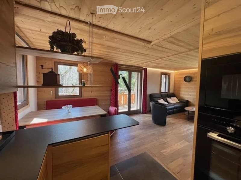 Open-plan living and dining area with wooden ceilings and floors, a black leather sofa, a dining table with chairs, and a modern oven.