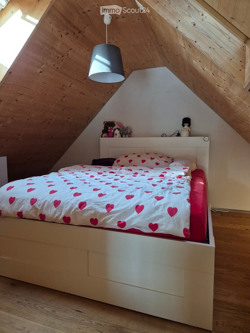 Cozy attic bedroom with wooden walls and slanted ceilings, featuring a white bed with heart-patterned bedding, a hanging lamp, and stuffed animals on the headboard.