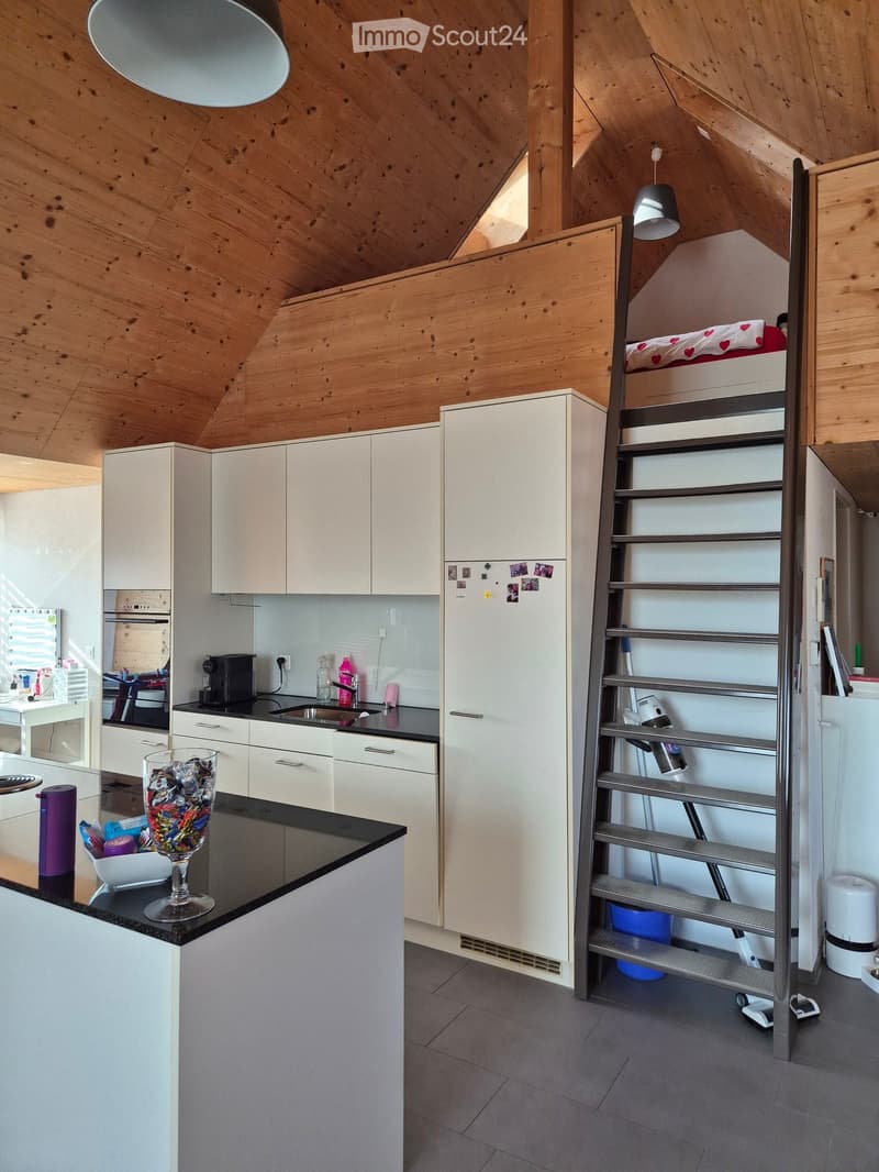 Modern kitchen with white cabinets, black countertops, and a staircase leading to a lofted bedroom area with a bed and lamp.