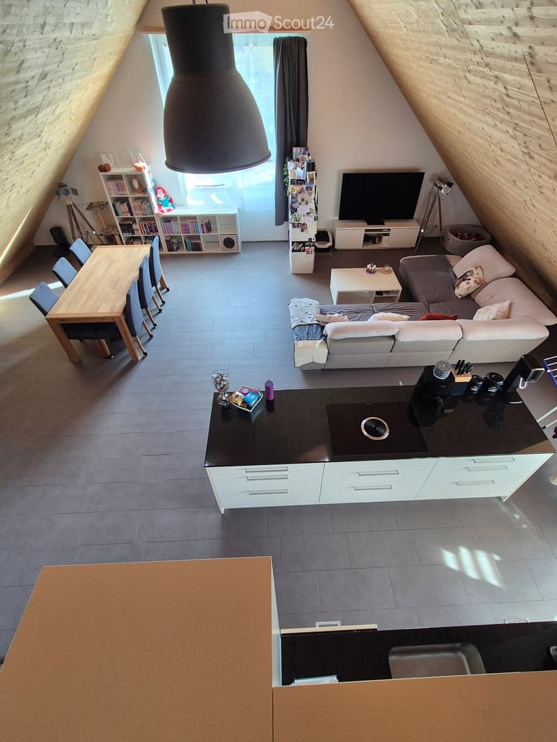 Open-plan living and dining area with a modern kitchen island, sofa, coffee table, dining table, and chairs. The space features exposed wooden beams and large windows allowing natural light.