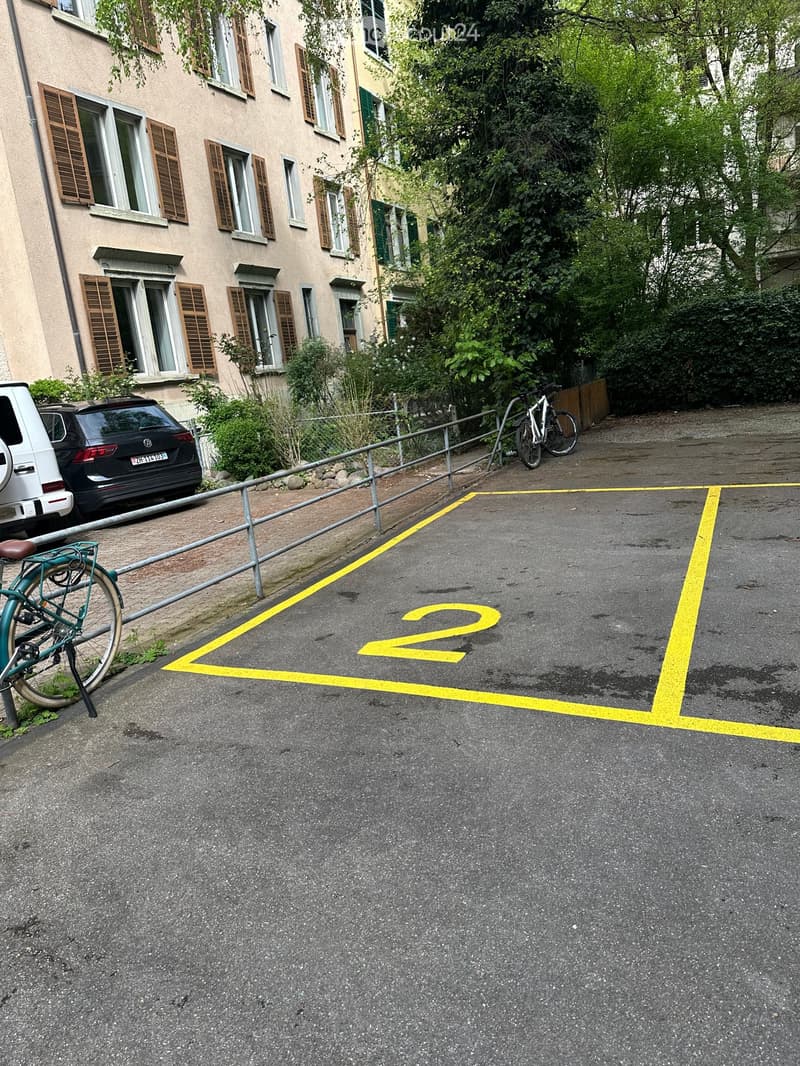 Multi-story residential building with multiple windows and shutters, adjacent to a parking area with marked spaces and bicycles.