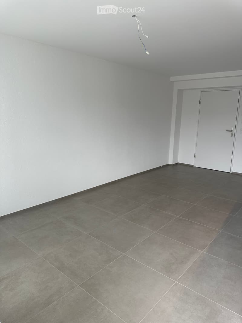 Empty room with grey tiled floor and white walls, featuring a white door and electrical outlet visible on the wall.