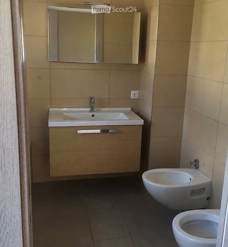 Modern bathroom with beige tiles, a white sink with a mirror above, and a toilet and bidet below.